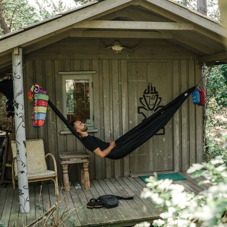 Load image into Gallery viewer, Person relaxing in a black hammock outside a wooden cabin in a forest setting.
